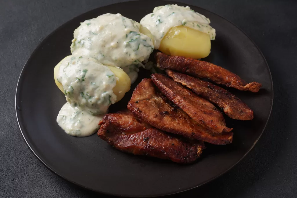 Stegt Flæsk med Persillesovs são pedaços crocantes de barriga de porco fritos com um molho cremoso de salsa, acompanhados de batatas cozidas.