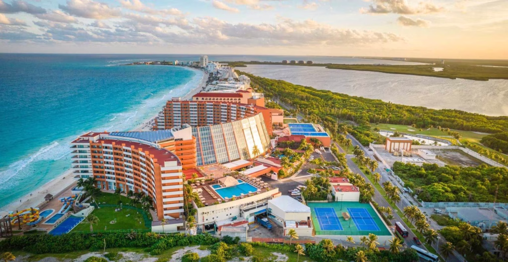 Vista aérea do Crown Paradise Club Cancún, com prédios de frente para o mar azul-turquesa, piscinas e escorregadores na areia.