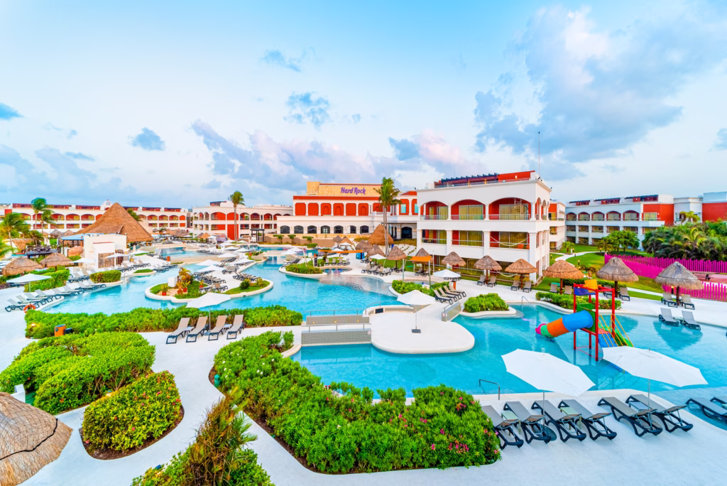 Piscinas do Hard Rock Hotel Riviera Maya cercadas por jardins, com área infantil de escorregador e prédios em tons de vermelho e branco.