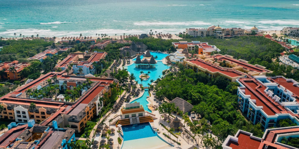 Vista aérea do Iberostar Selection Paraíso Maya, com piscinas entre jardins, telhados de palha e o mar do Caribe ao fundo.